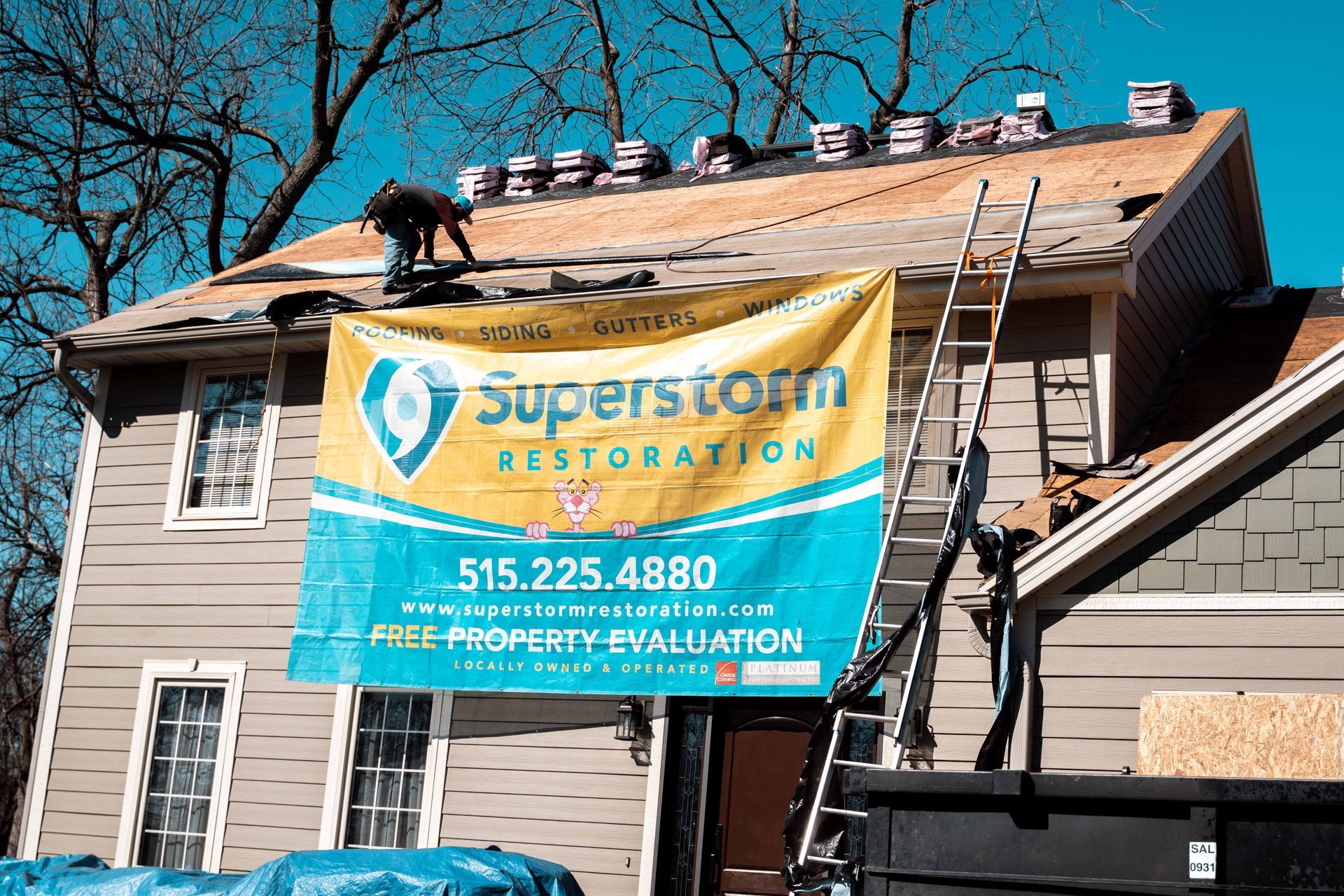 Man on roof installing shingles, Superstorm Restoration banner on house. Ladder, dumpster visible.
