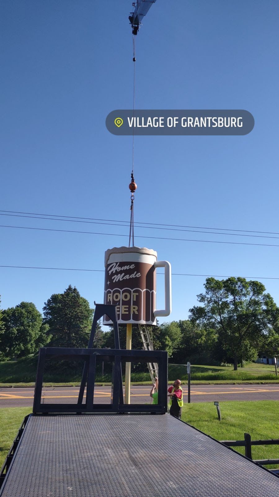 A large coffee mug sign is being lifted by a crane in the Village of Grantsburg