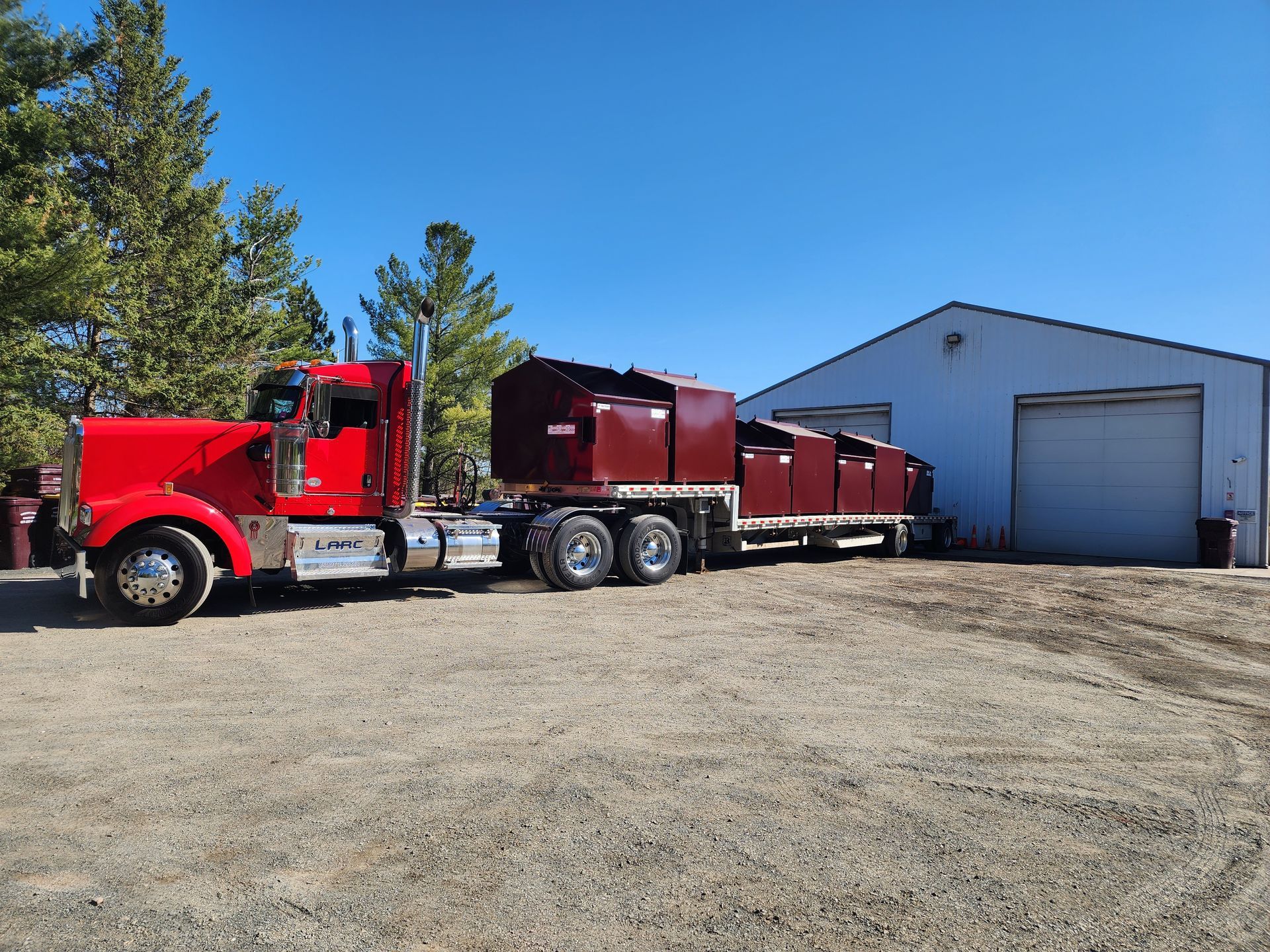 Red semi-truck with maroon trailer parked beside a white warehouse and garage door under a blue sky