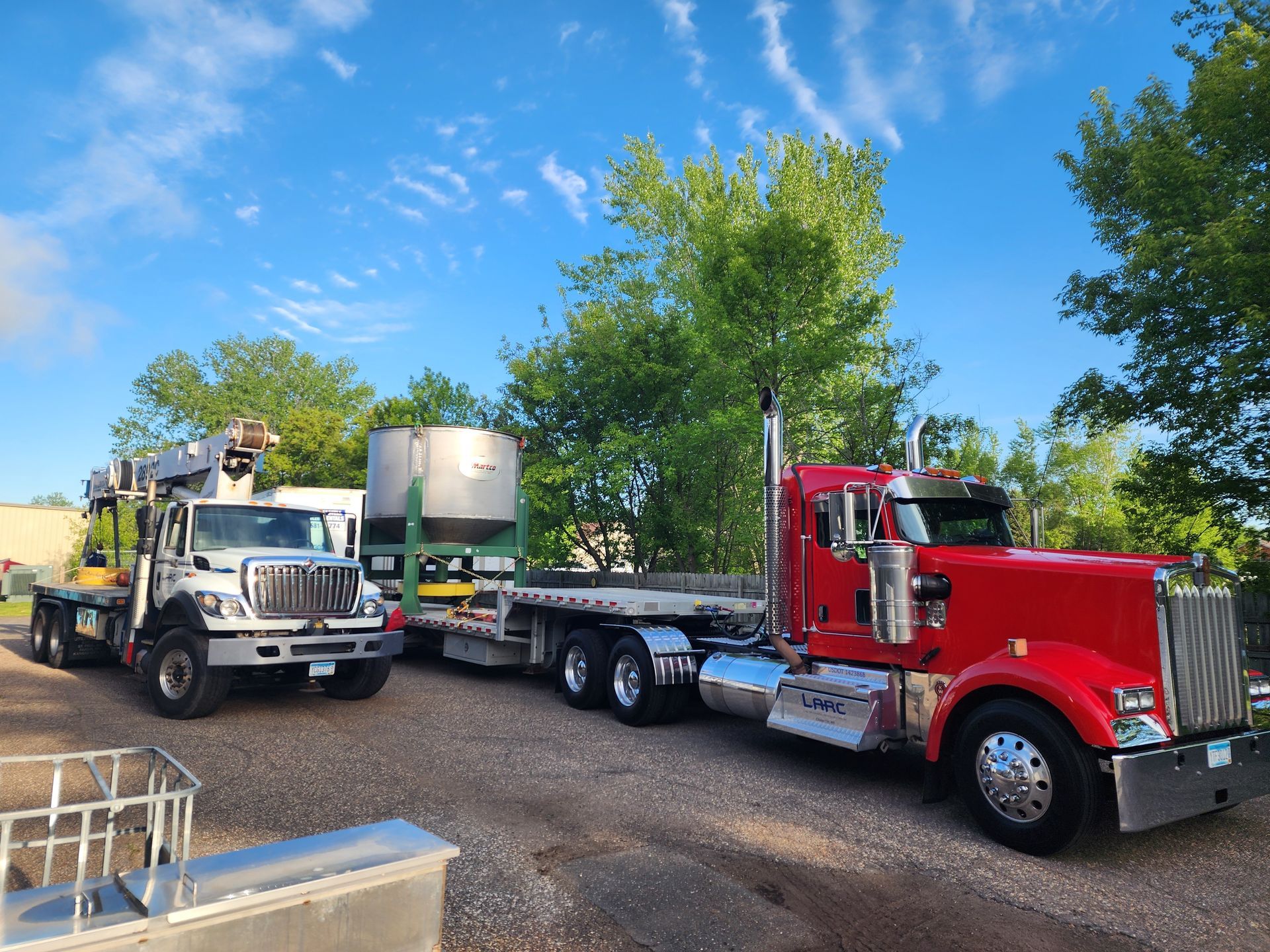 Red semi truck towing a flatbed with a white truck, parked outdoors under a blue sky.