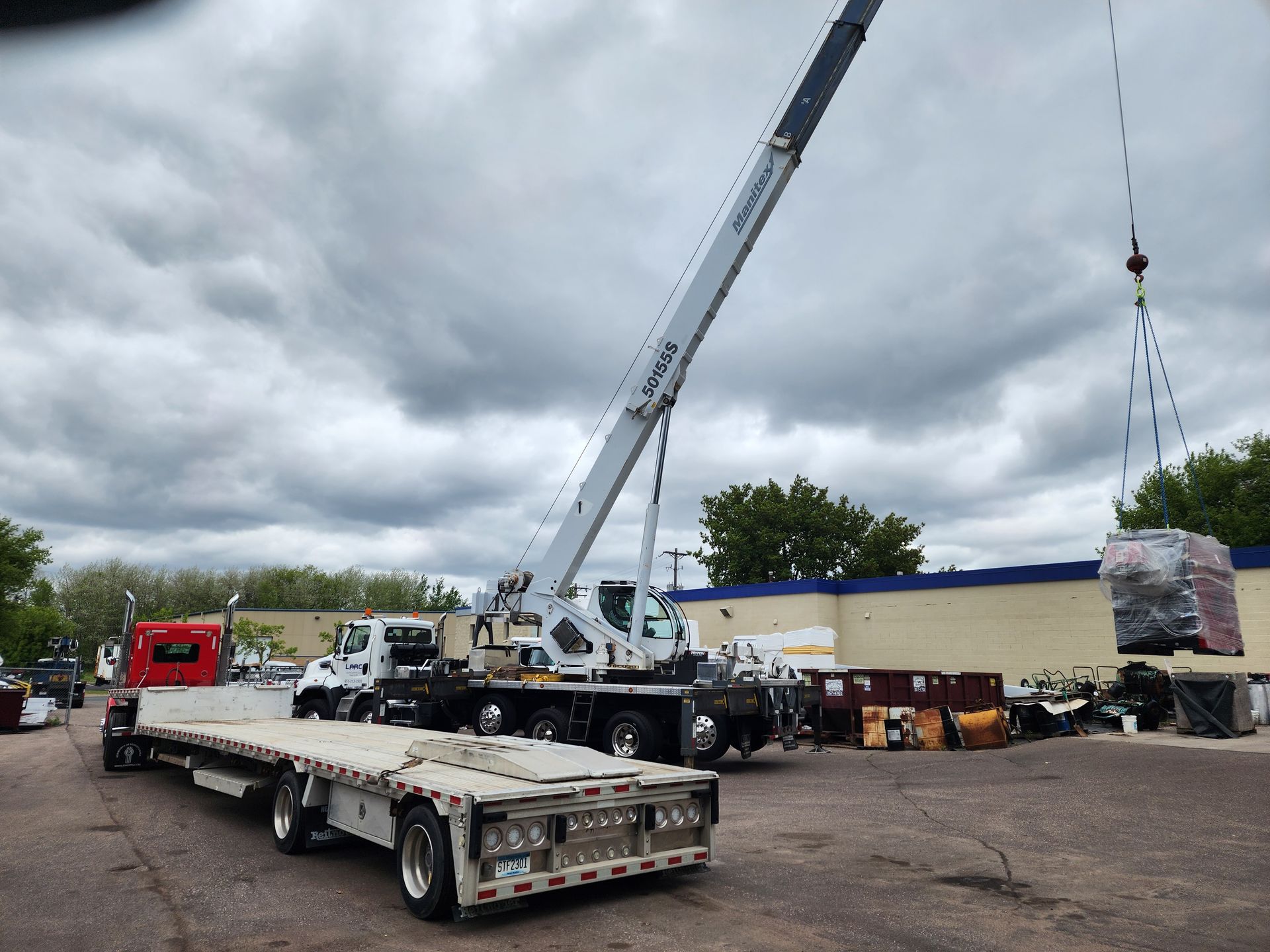 Flatbed crane truck lifting a wrapped pallet in a lot under cloudy skies