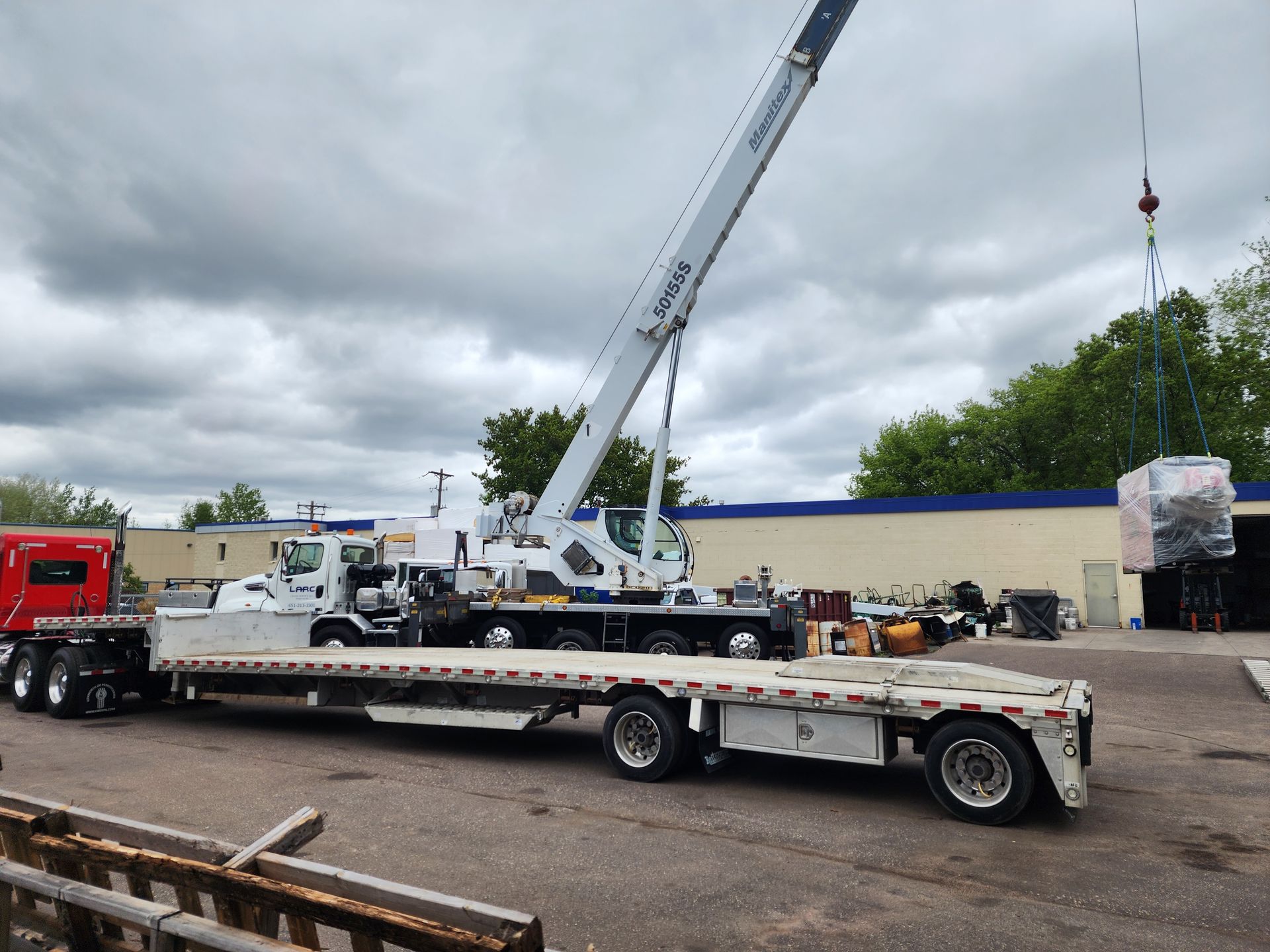 Crane loading equipment onto a flatbed trailer in an industrial yard under cloudy skies
