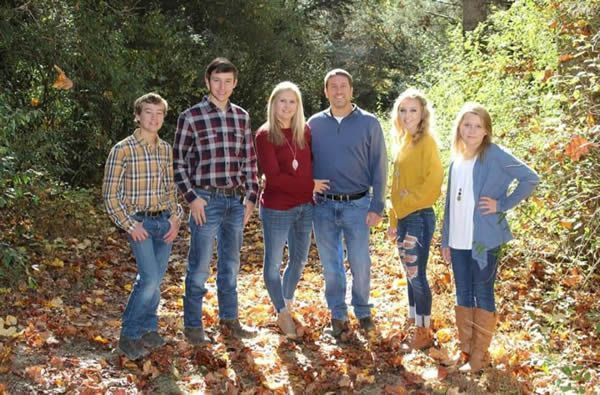 Family of six poses outdoors in autumn; foliage surrounds them.