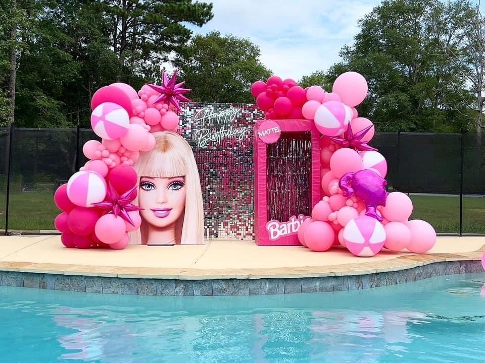 Barbie-themed pool party decorations: pink balloon arches, a Barbie head, and a box cutout, near a pool.