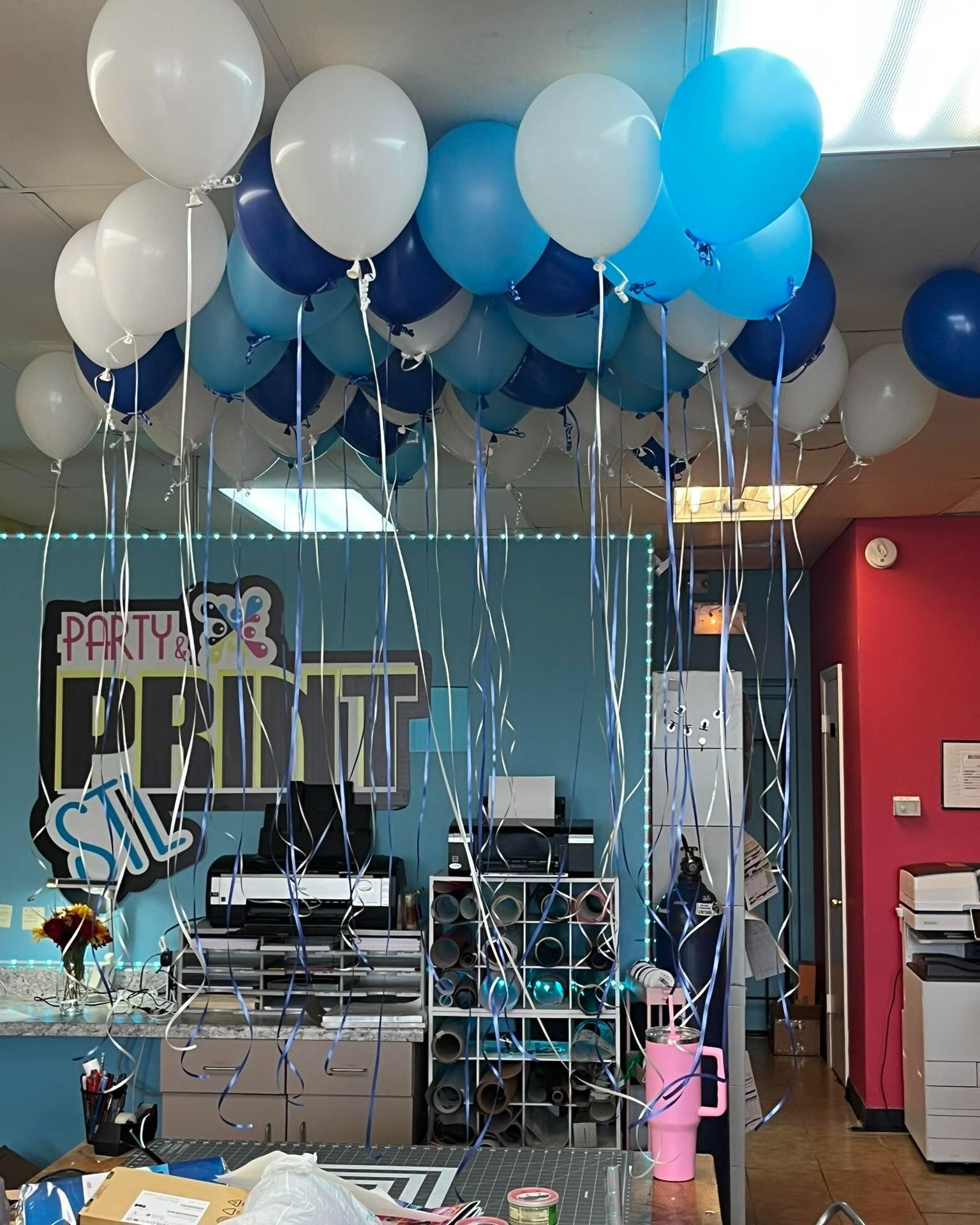 Blue and white balloons decorate a printing shop, with some balloons attached to the ceiling and some with streamers.