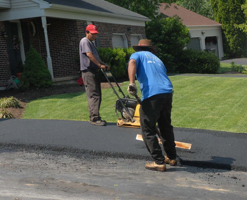 Two workers paving a driveway with a compactor machine on a sunny day.