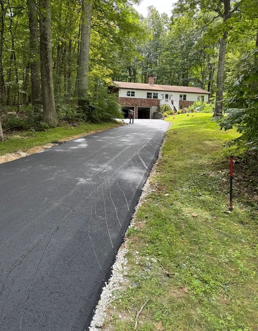 Newly paved black asphalt driveway leading to a house in a wooded area.