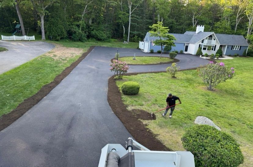 Black asphalt driveway curves to a light blue house. A person edges grass. Lush green lawn with trees.