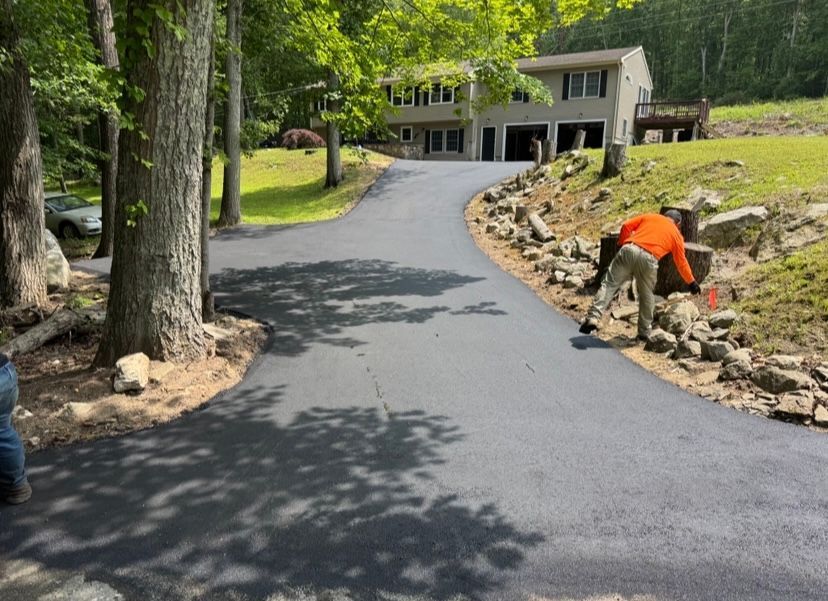 Newly paved driveway leading to a two-story house, worker placing a wooden beam next to rocks.