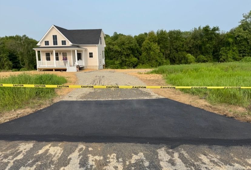 A newly paved driveway leads to a small, light-colored house. Yellow caution tape blocks access.
