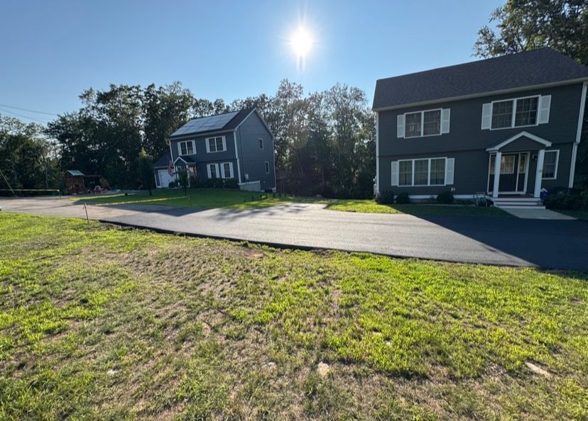 Two-story houses with dark siding and white trim on a sunny day. A paved road and grassy area in the foreground.