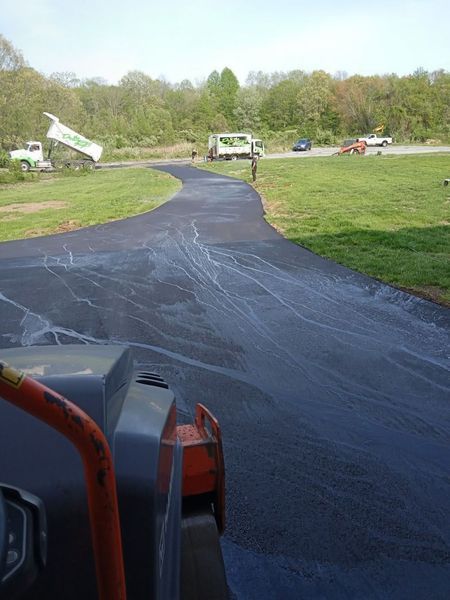 Asphalt driveway being rolled by a machine with trucks and vehicles in the background. Green grass on either side.