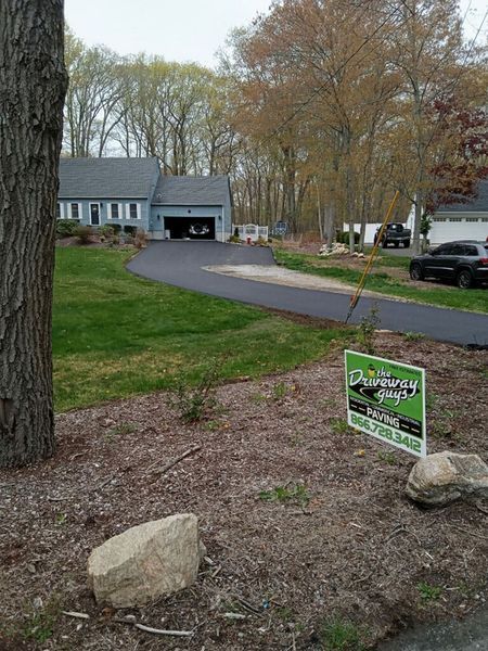 A house with a newly paved driveway; lawn and trees surround the home. A sign for