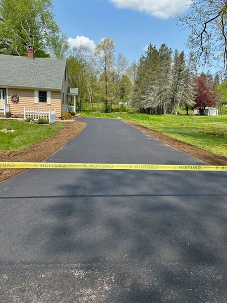 Freshly paved asphalt driveway with caution tape, leading to a house in a green, sunny setting.