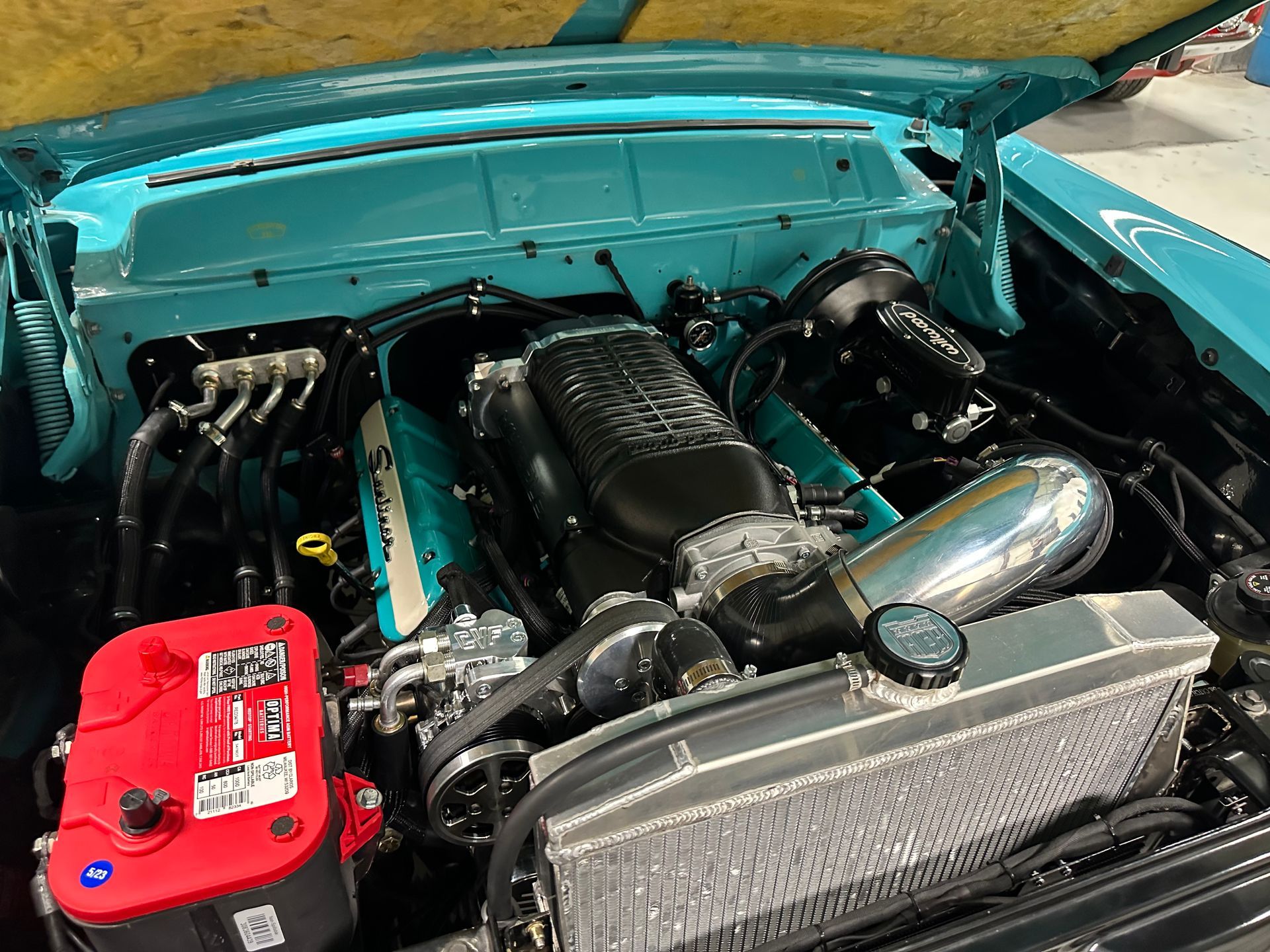 The engine of a blue car with the hood up and a red battery.