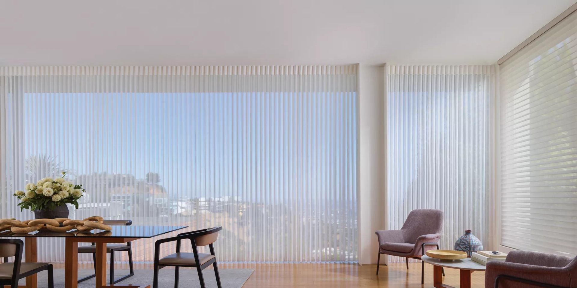 Bright modern living room with sheer curtains, wooden table, chairs, and a city view through large windows