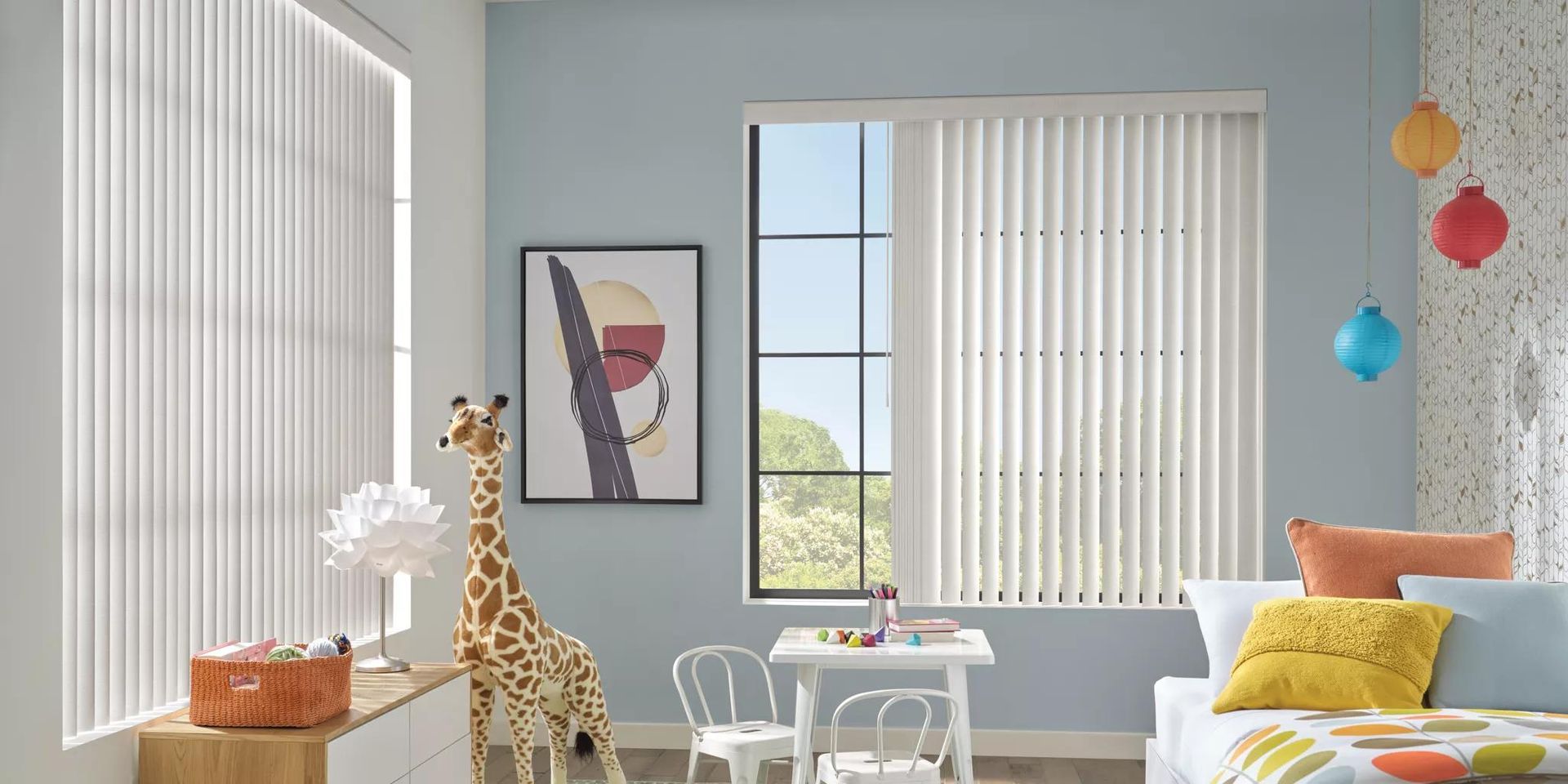 Bright nursery with a giraffe toy, yellow couch, colorful hanging balls, and a framed wall print by the window.