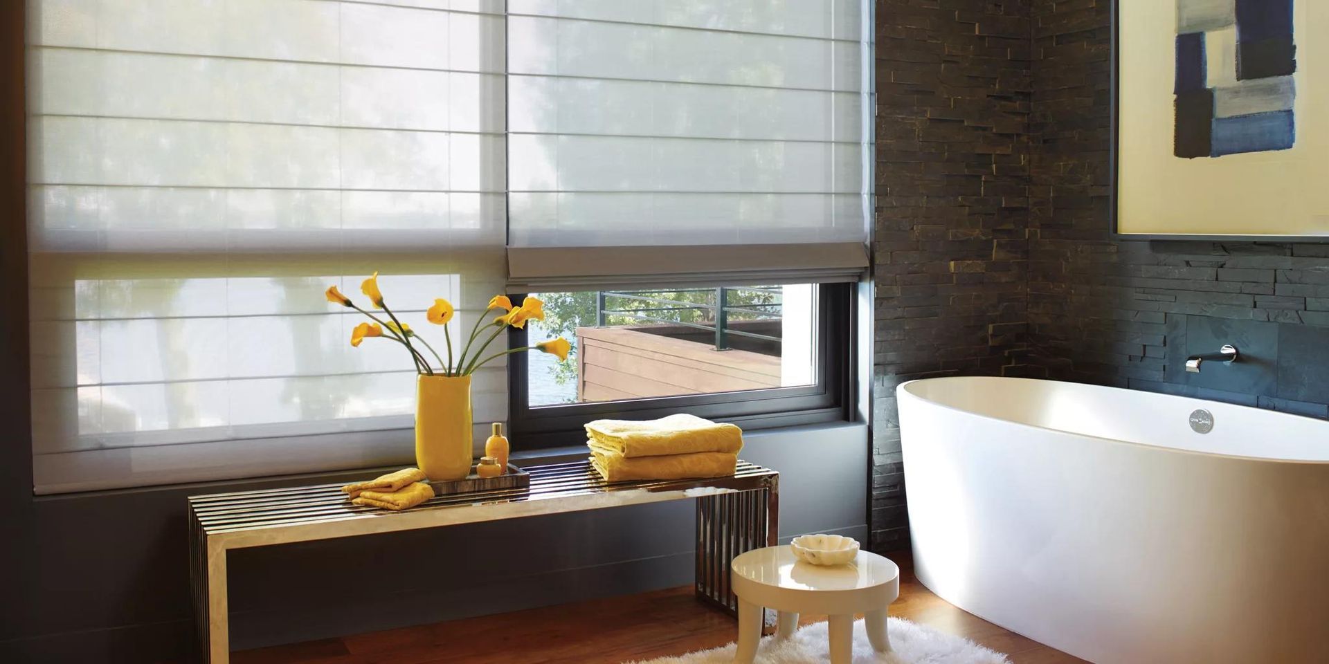 Modern bathroom with a white tub, dark tile wall, window blinds, and a wooden bench with towels and a vase of flowers