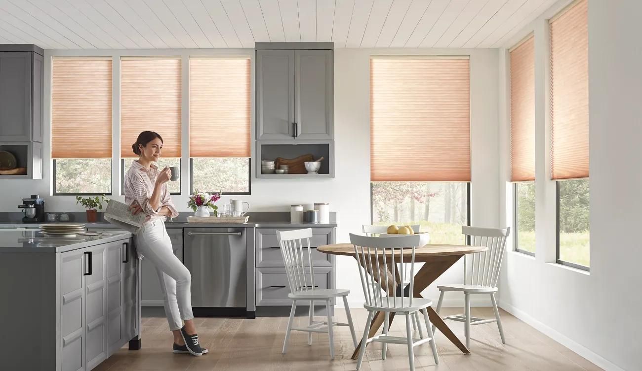 Bright kitchen with a person standing by the counter beside a small dining table and windows with blinds.