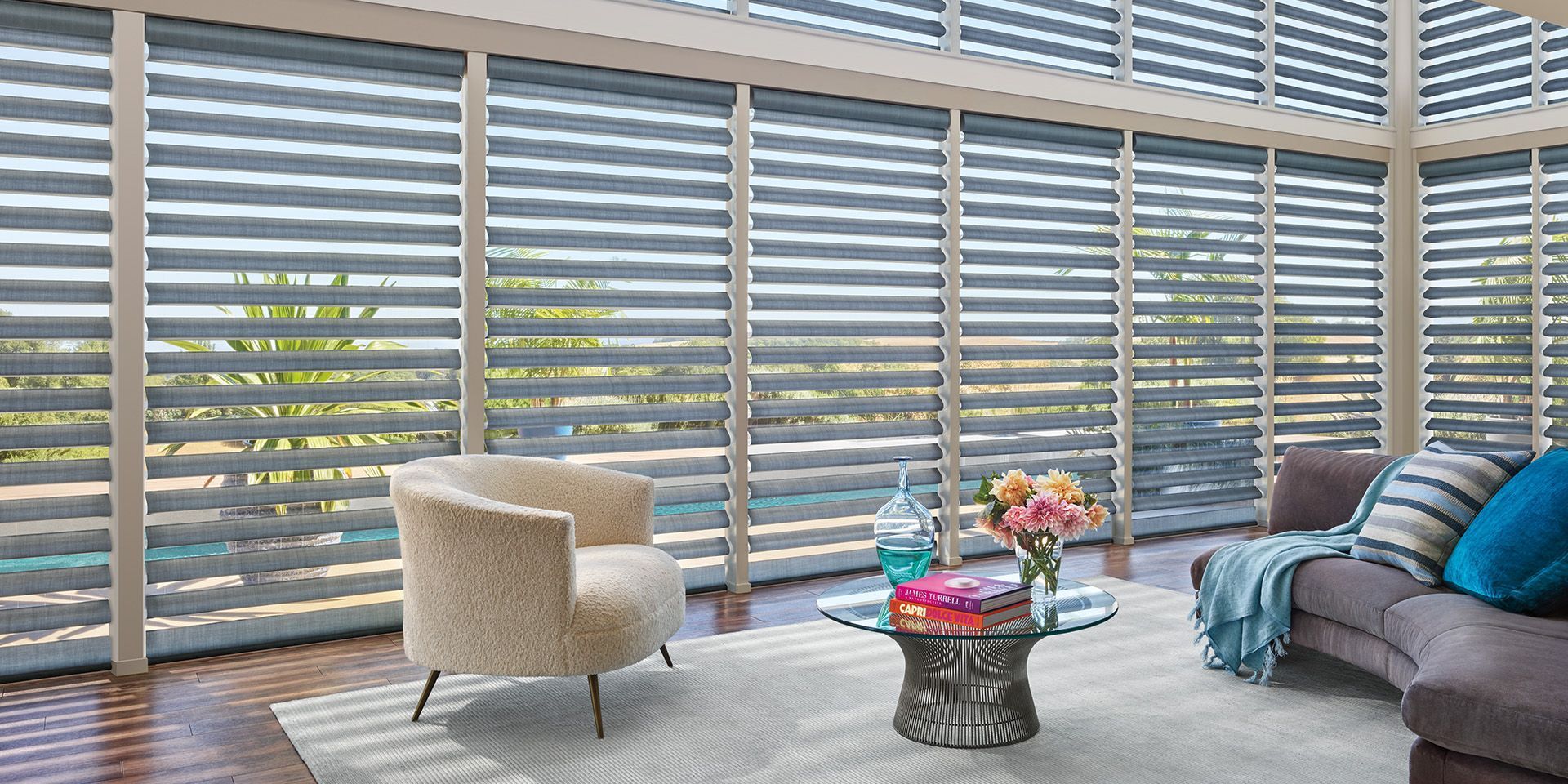 Modern living room with striped blinds, gray sofa, beige chair, and small table with flowers and decor