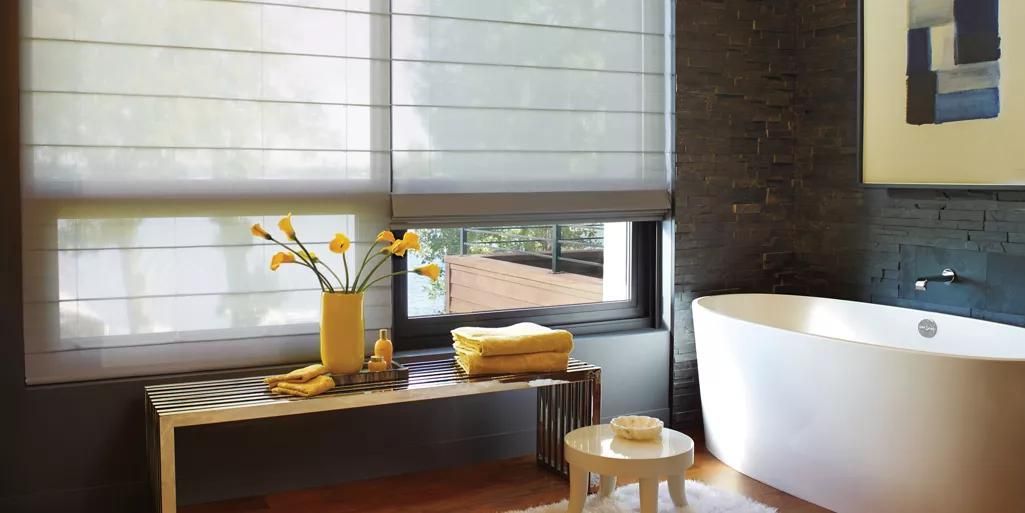 Modern bathroom with a freestanding tub, vanity, and window with blinds, plus flowers on a stool