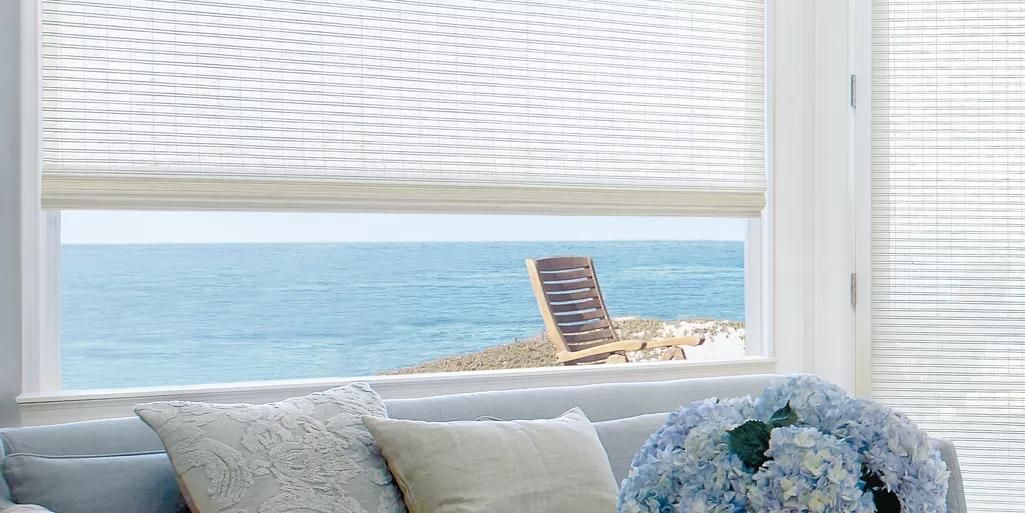 Coastal bedroom window with ocean view, white blinds, and a blue floral pillow on the bed