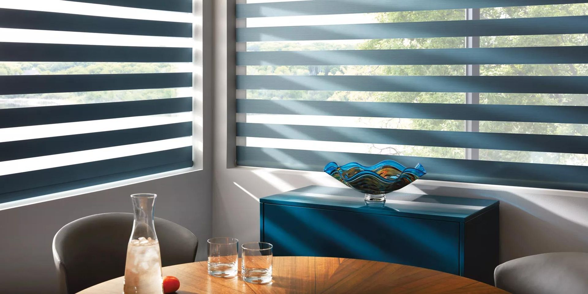 Dining table by window blinds with a glass bottle, glasses, and a blue table with a fruit bowl.