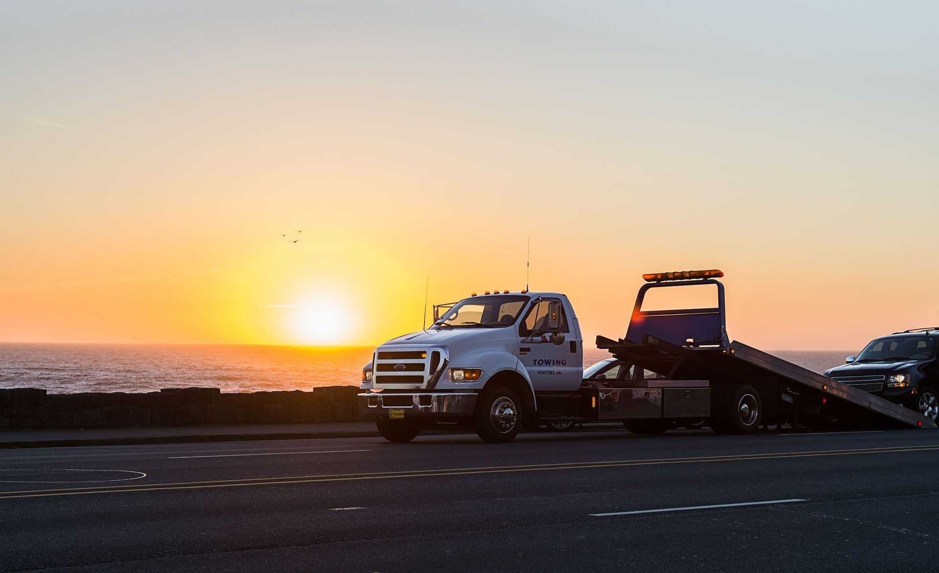 Tow truck towing a car near the ocean at sunset; orange sky, water.