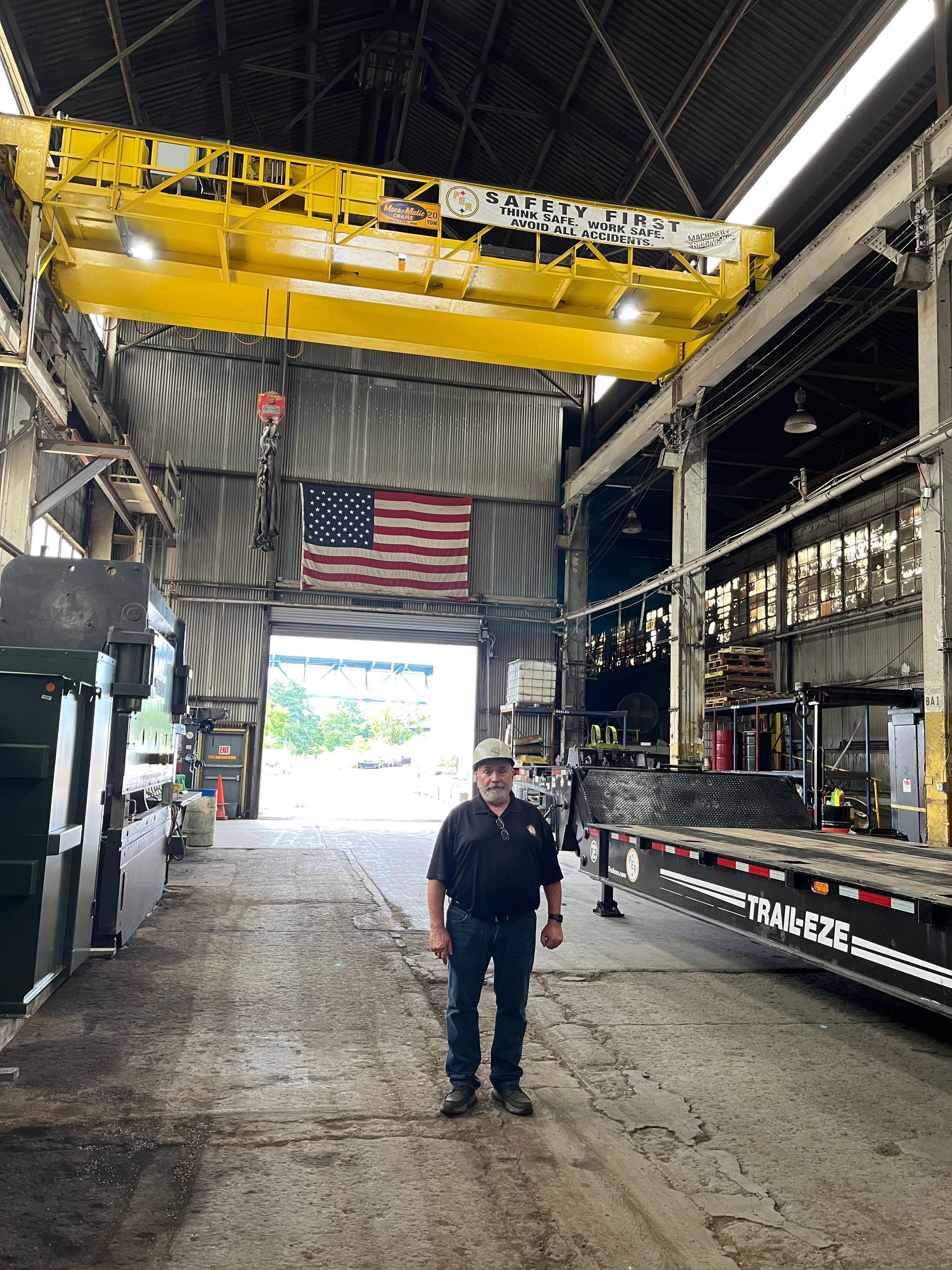 A man is standing in front of a large yellow crane in a factory.