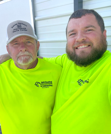 Two smiling workers in neon yellow shirts and caps posing outdoors by a white building.