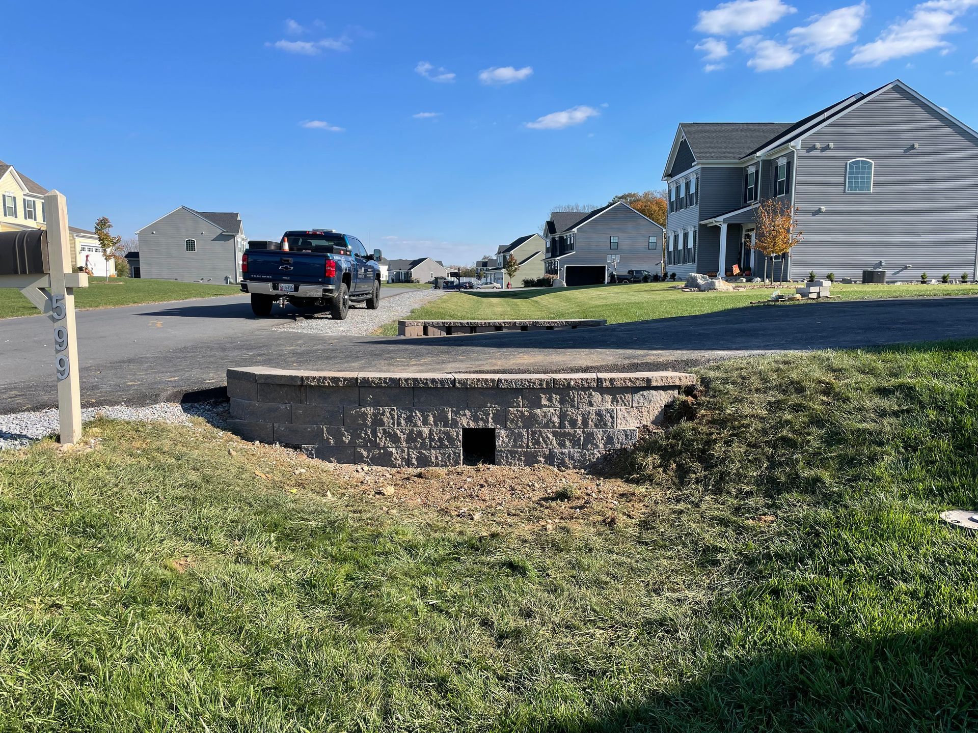 A stone drainage headwall sits in a grassy yard in front of a residential street with houses and a parked blue truck.