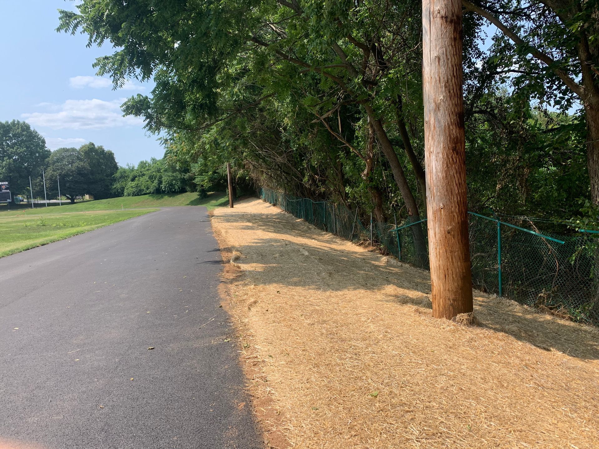 A paved trail runs next to a wood-mulched path lined by a chain-link fence, green trees, and a wooden utility pole.
