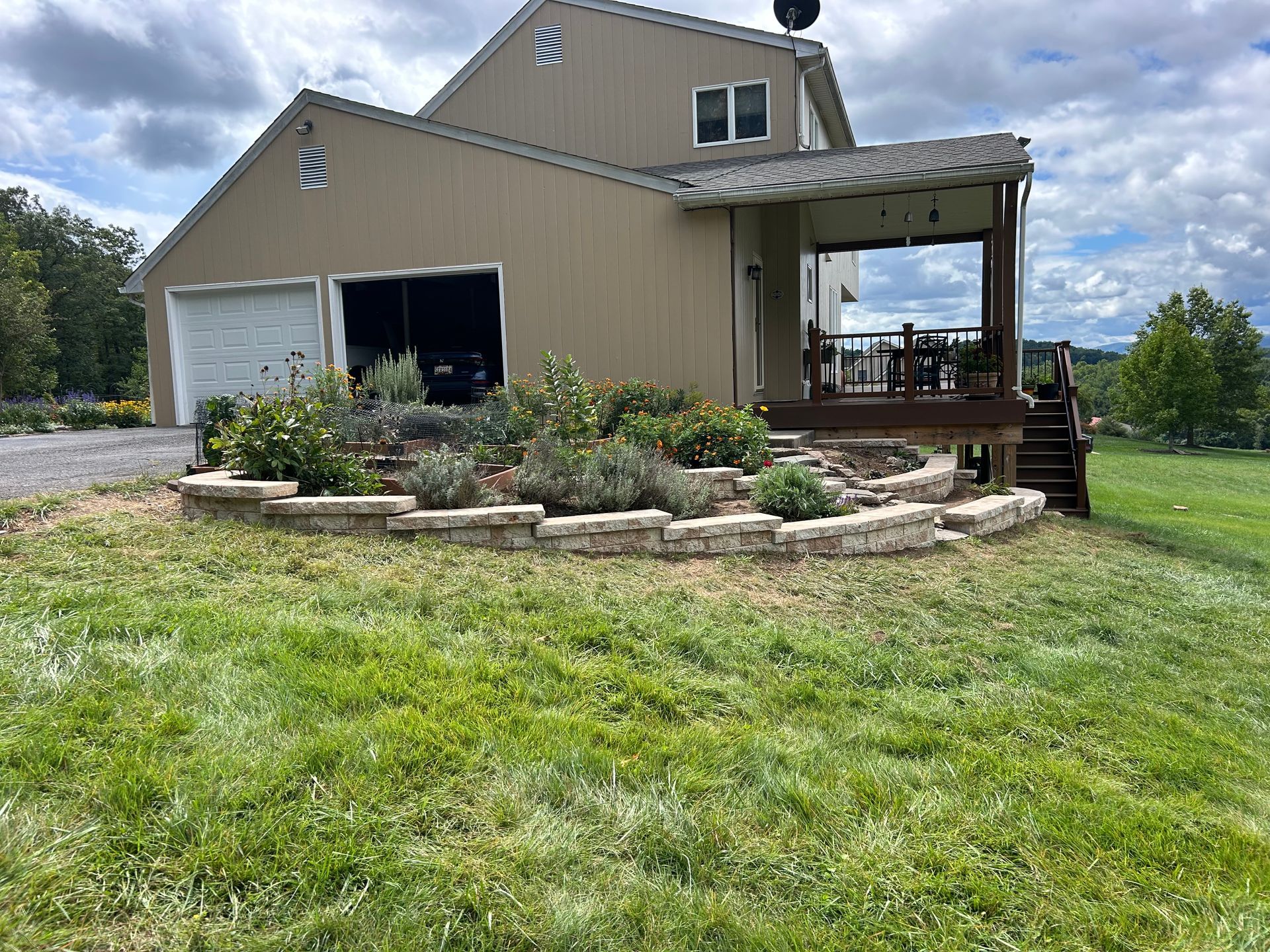 A tan two-story house with a white garage door, a wooden porch, and a stone-walled garden bed in the grassy yard.