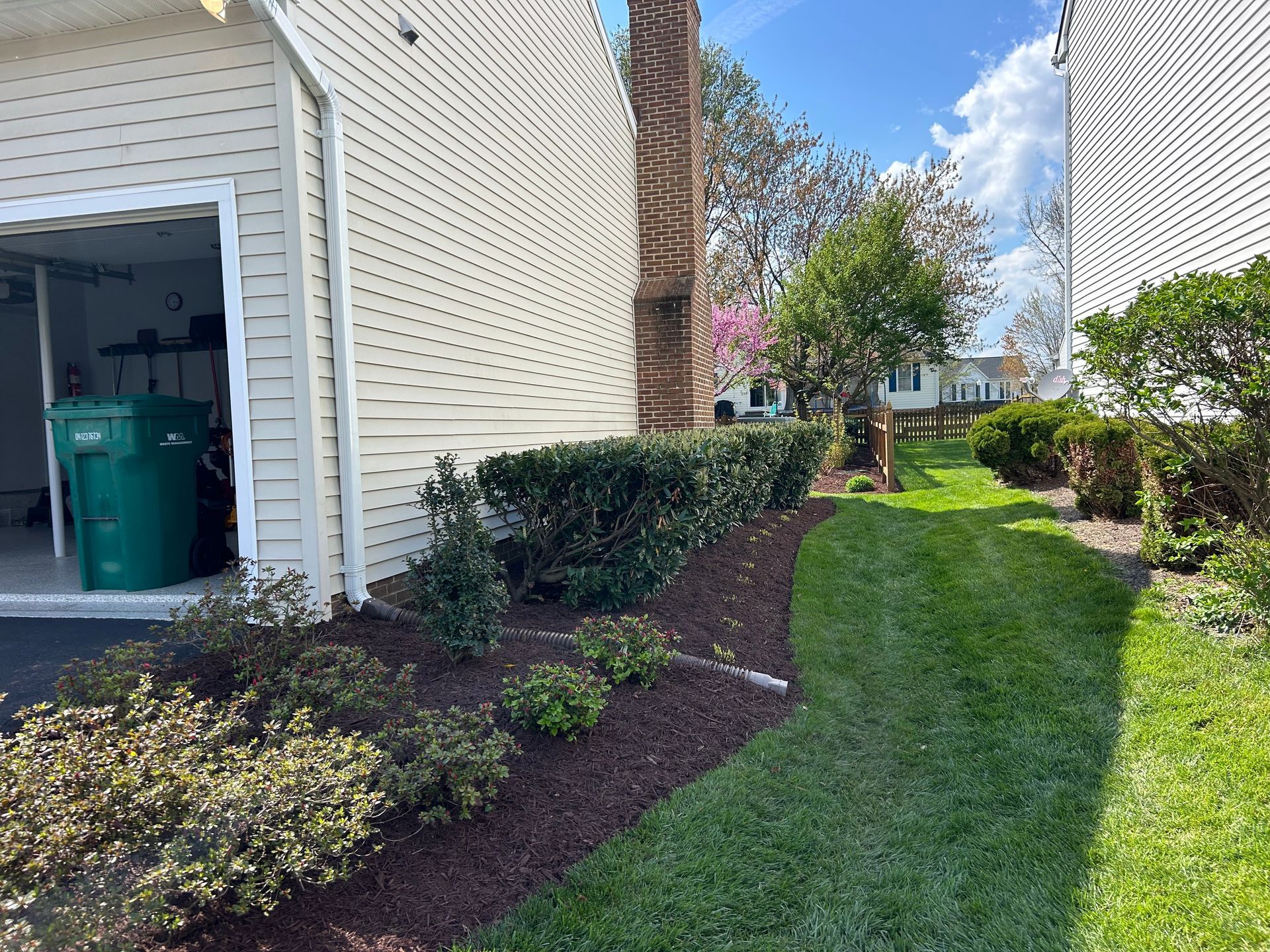 A beige house with an open garage and a green recycling bin, alongside a mulched garden bed and a grassy yard.