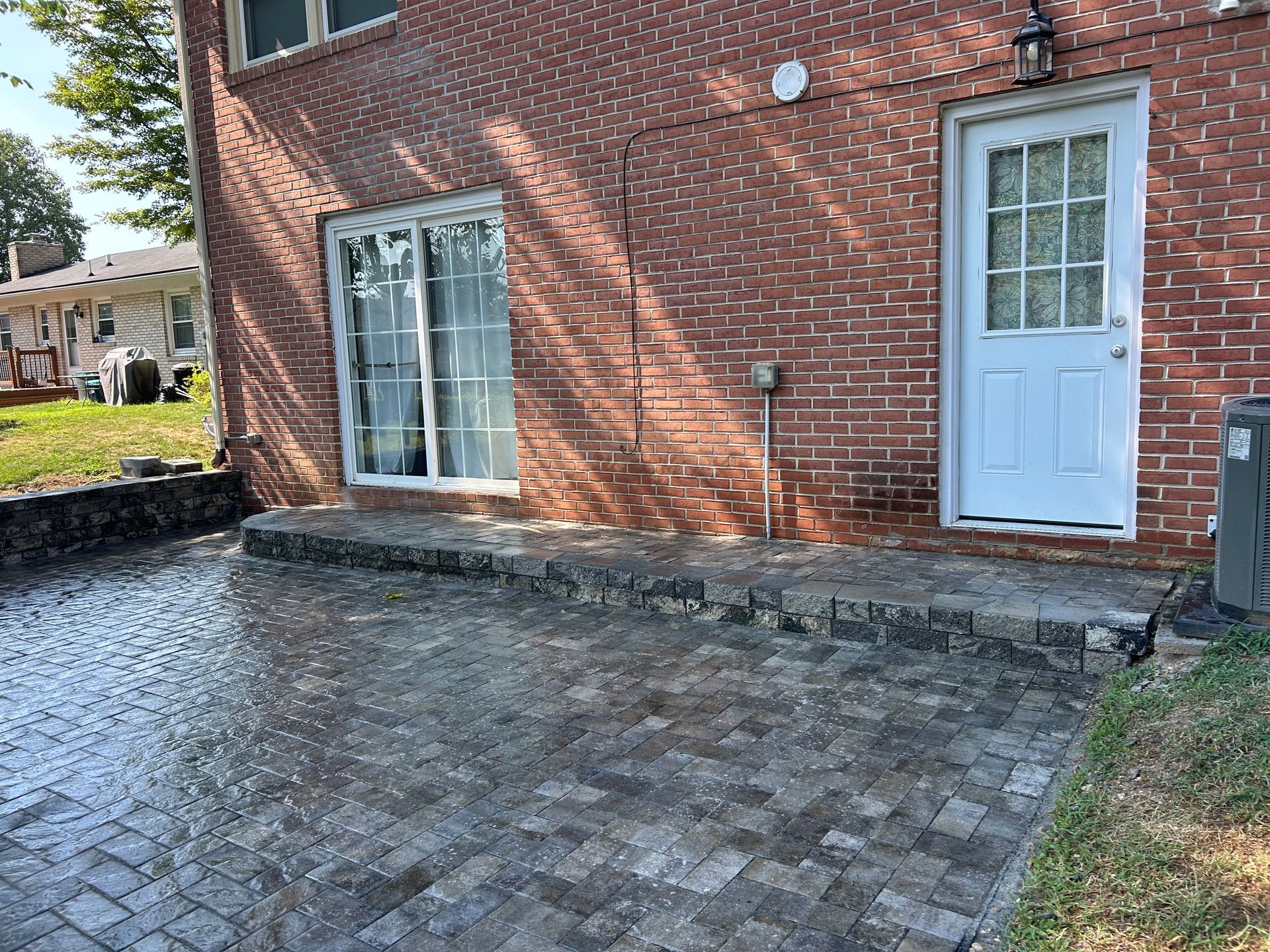 A stone patio with a low retaining wall sits against a red brick house featuring a white sliding door and a white door.