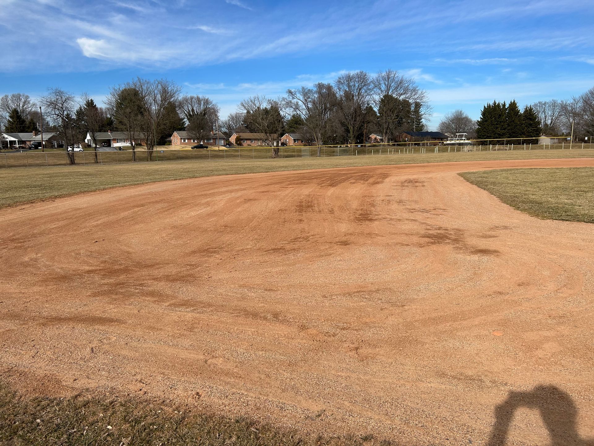 A dirt baseball infield viewed from the grass, with a residential neighborhood and bare trees in the background.