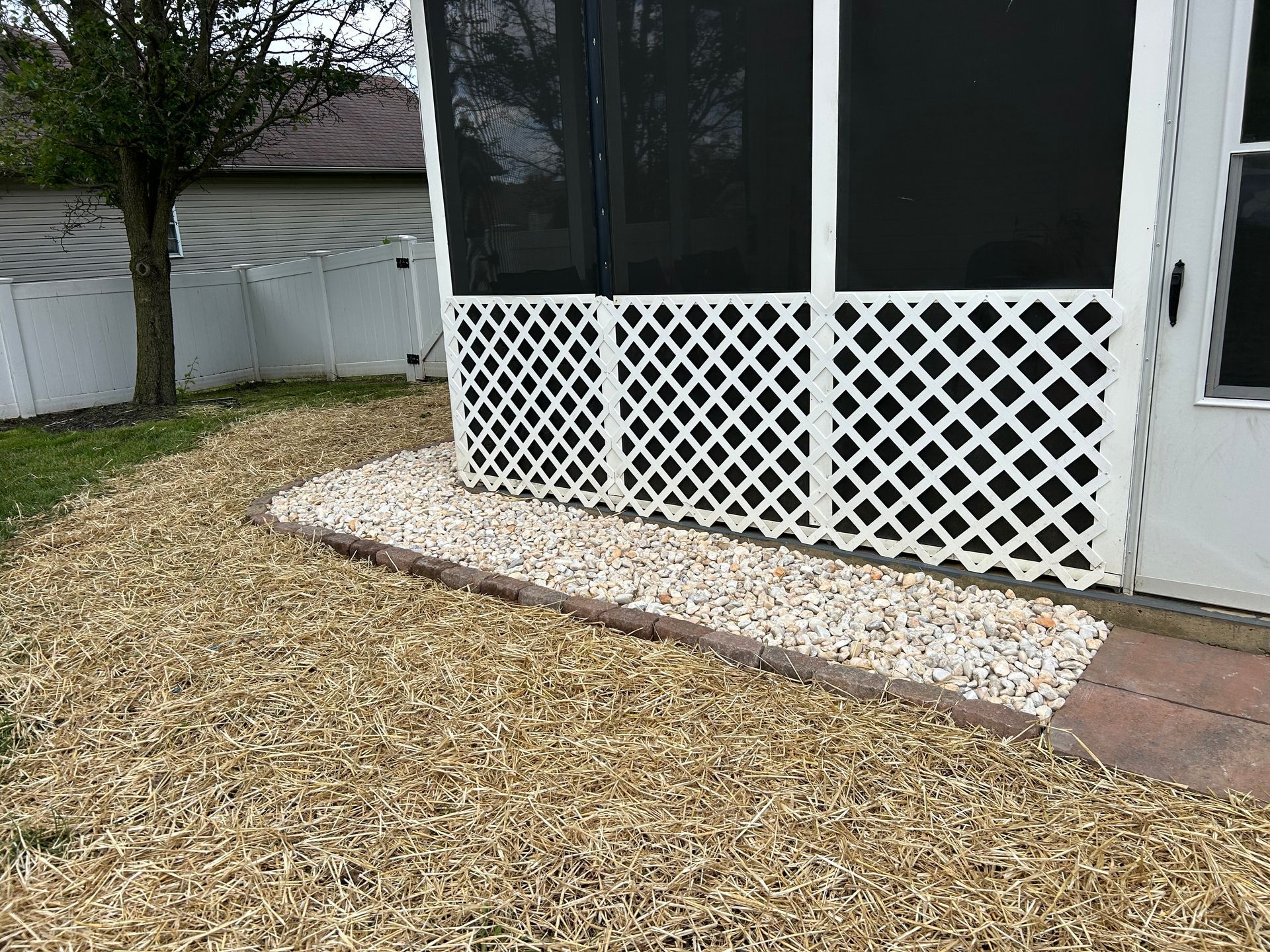 A white lattice skirt borders the base of a screened porch, flanked by light-colored gravel and wood chips on the lawn.