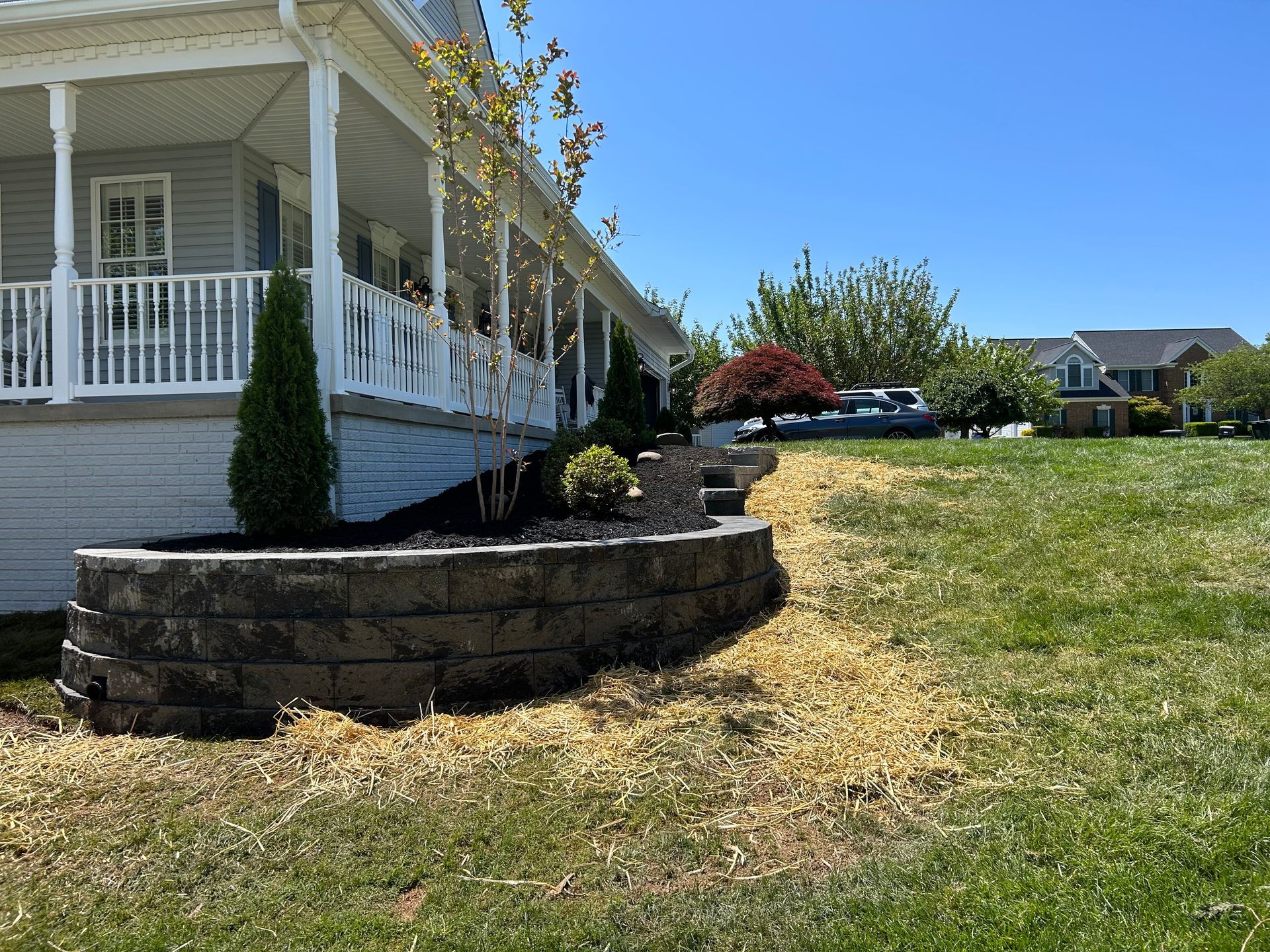 A retaining wall made of stacked grey stone blocks sits in front of a house porch with a lawn and trees in the background.
