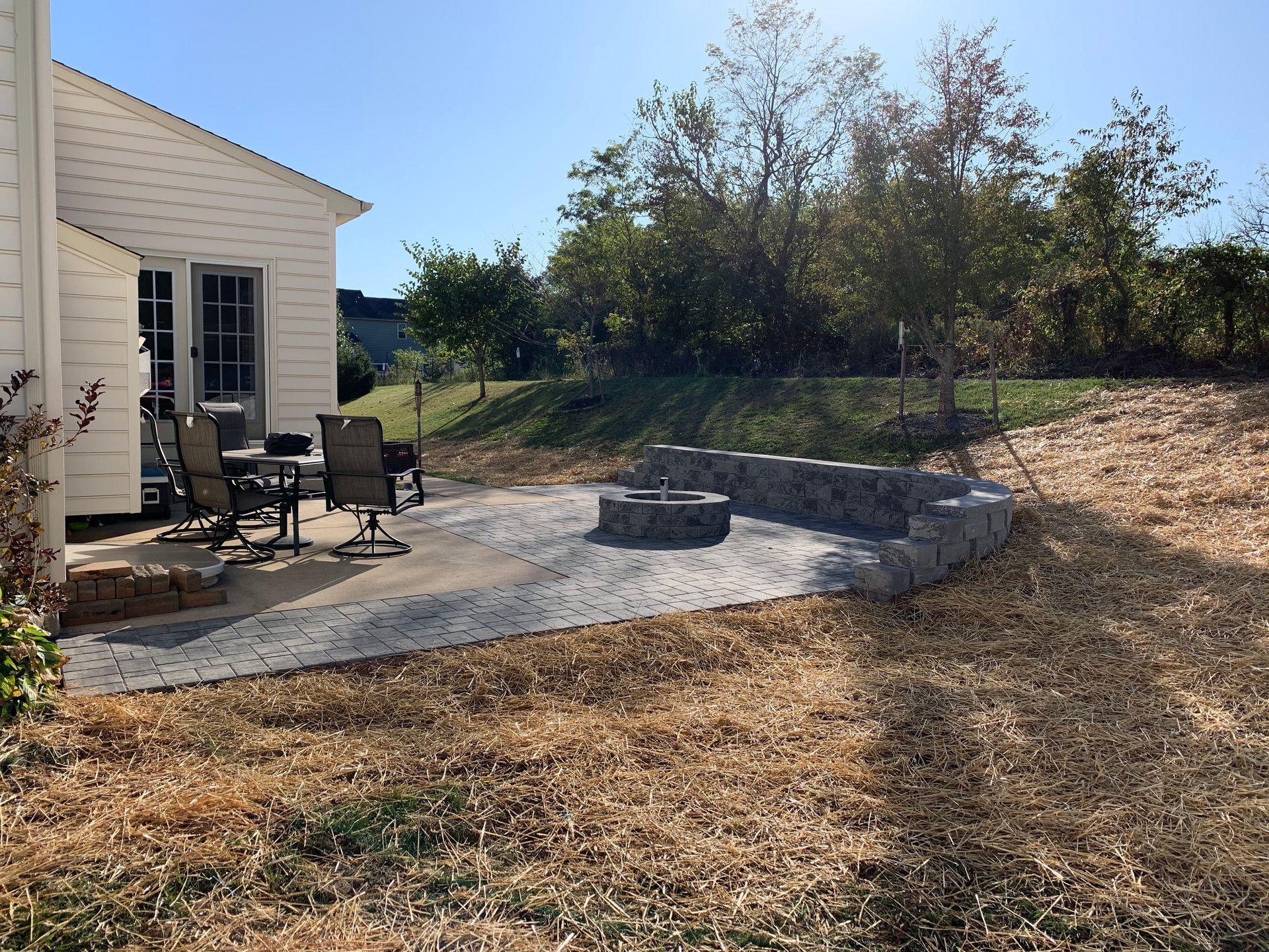 Patio with chairs and a fire pit next to a white house, surrounded by trees and a dry, grassy yard under a blue sky.