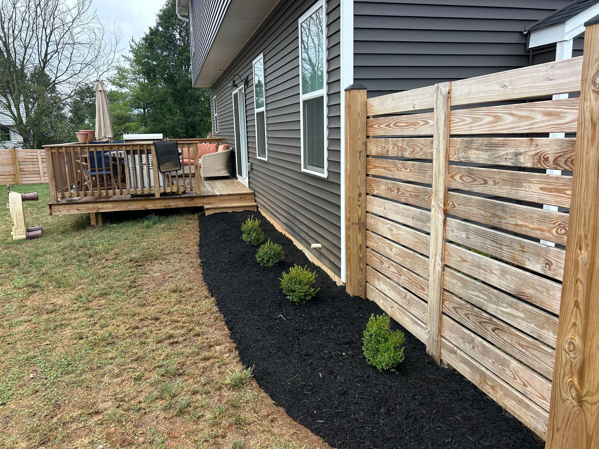 A horizontal wood slat fence runs along a house wall beside a garden bed filled with black mulch and small green shrubs.