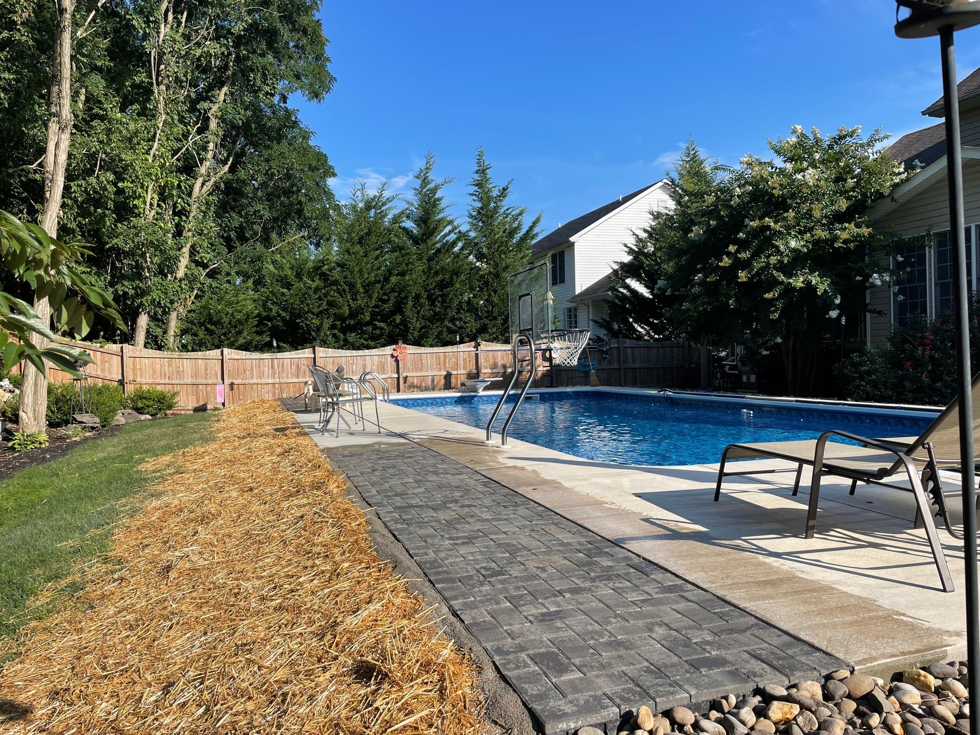 A bright, sunny backyard with an inground pool, stone patio, mulch bed, and a wooden privacy fence near trees.