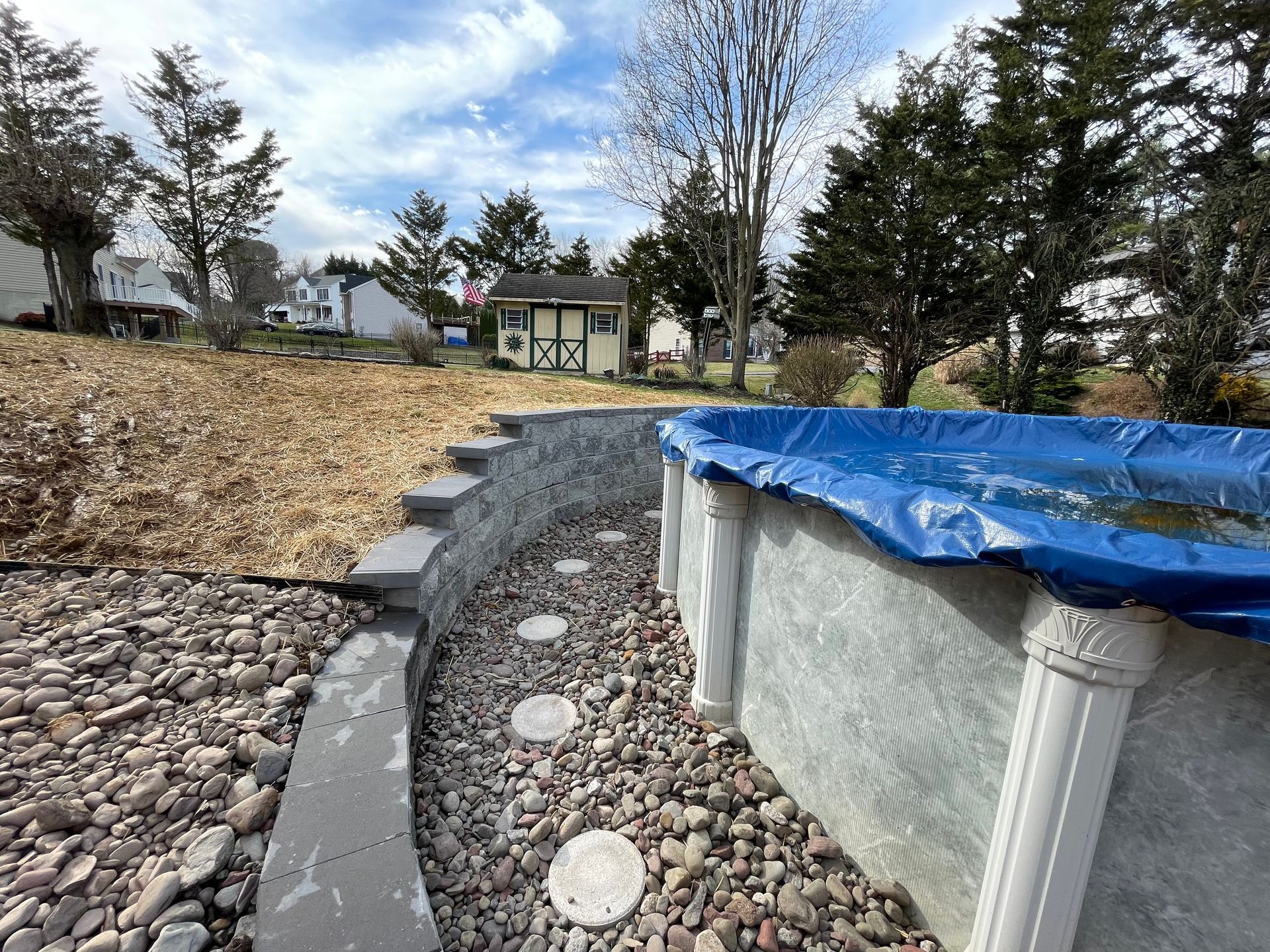 An above-ground pool with a blue cover, bordered by a stone retaining wall and a gravel path, with a shed in the distance.