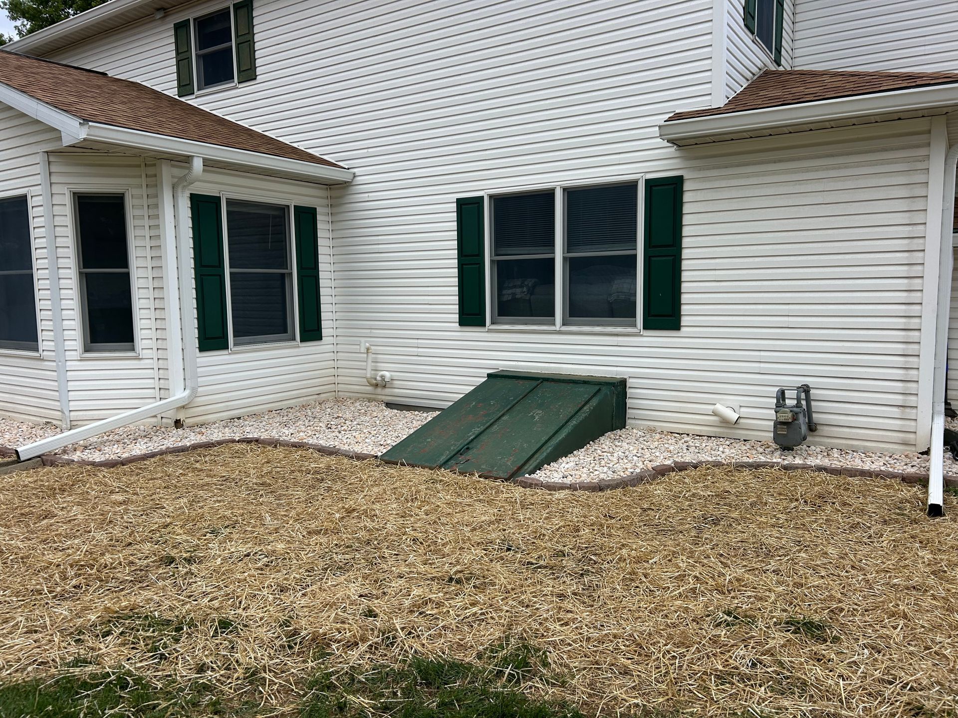White house exterior with dark green shutters, a slanted metal basement storm door, and a gravel-covered yard.