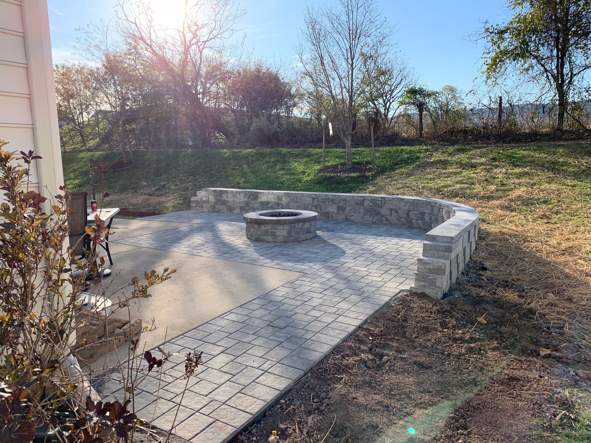 A stone patio with a central fire pit surrounded by a low curved stone retaining wall on a grassy backyard hillside.