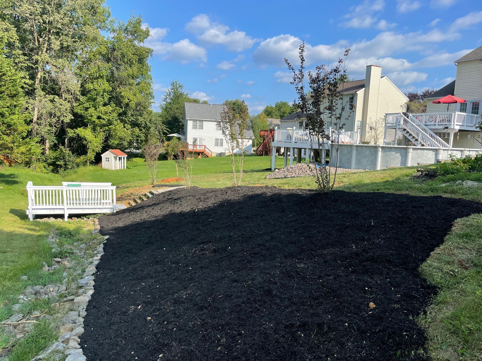 A backyard features a large, fresh patch of black mulch in the foreground, adjacent to a white wooden bridge and houses.