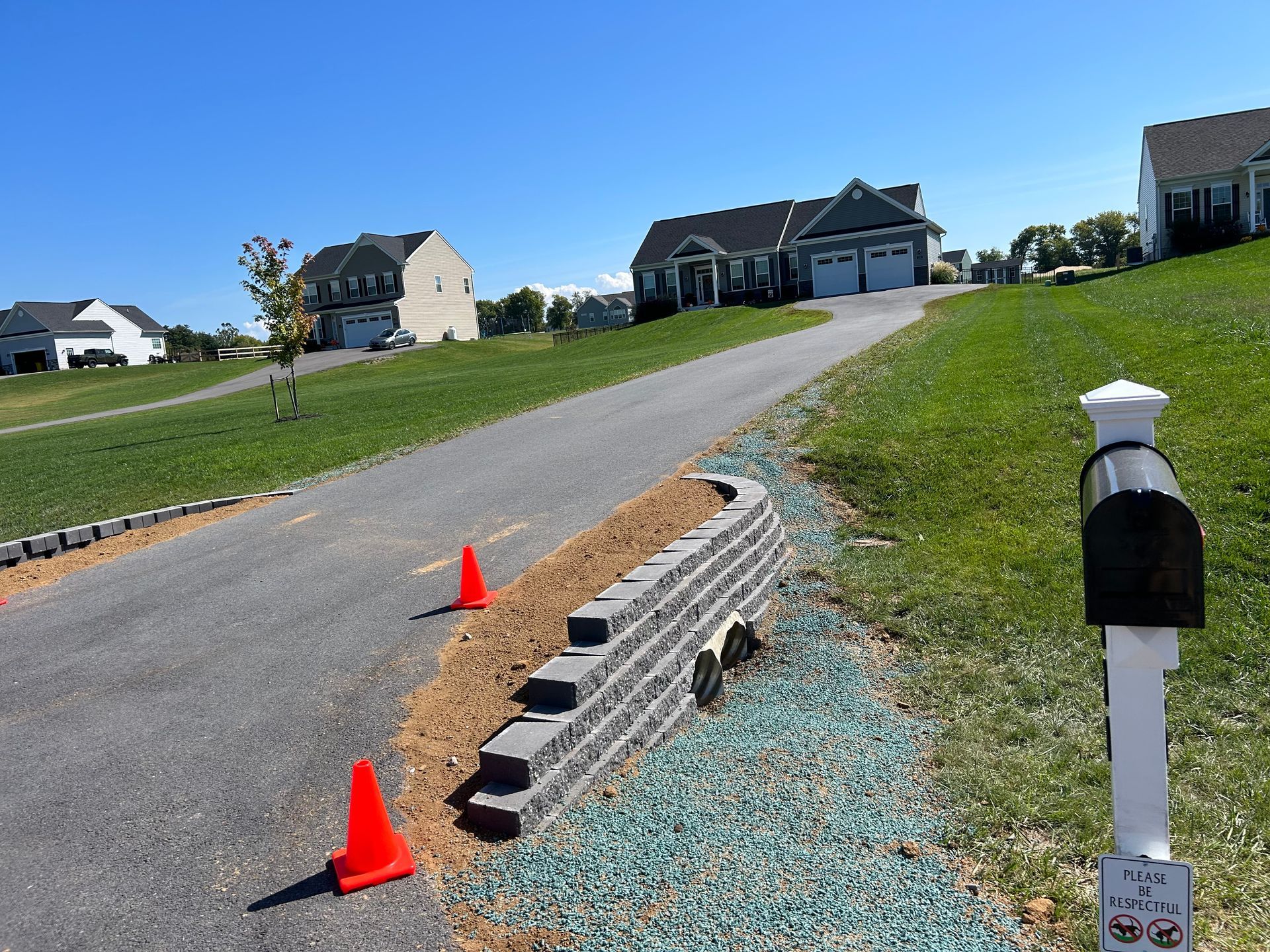 A gray stone retaining wall stands along a paved driveway with orange safety cones on a sunny day.