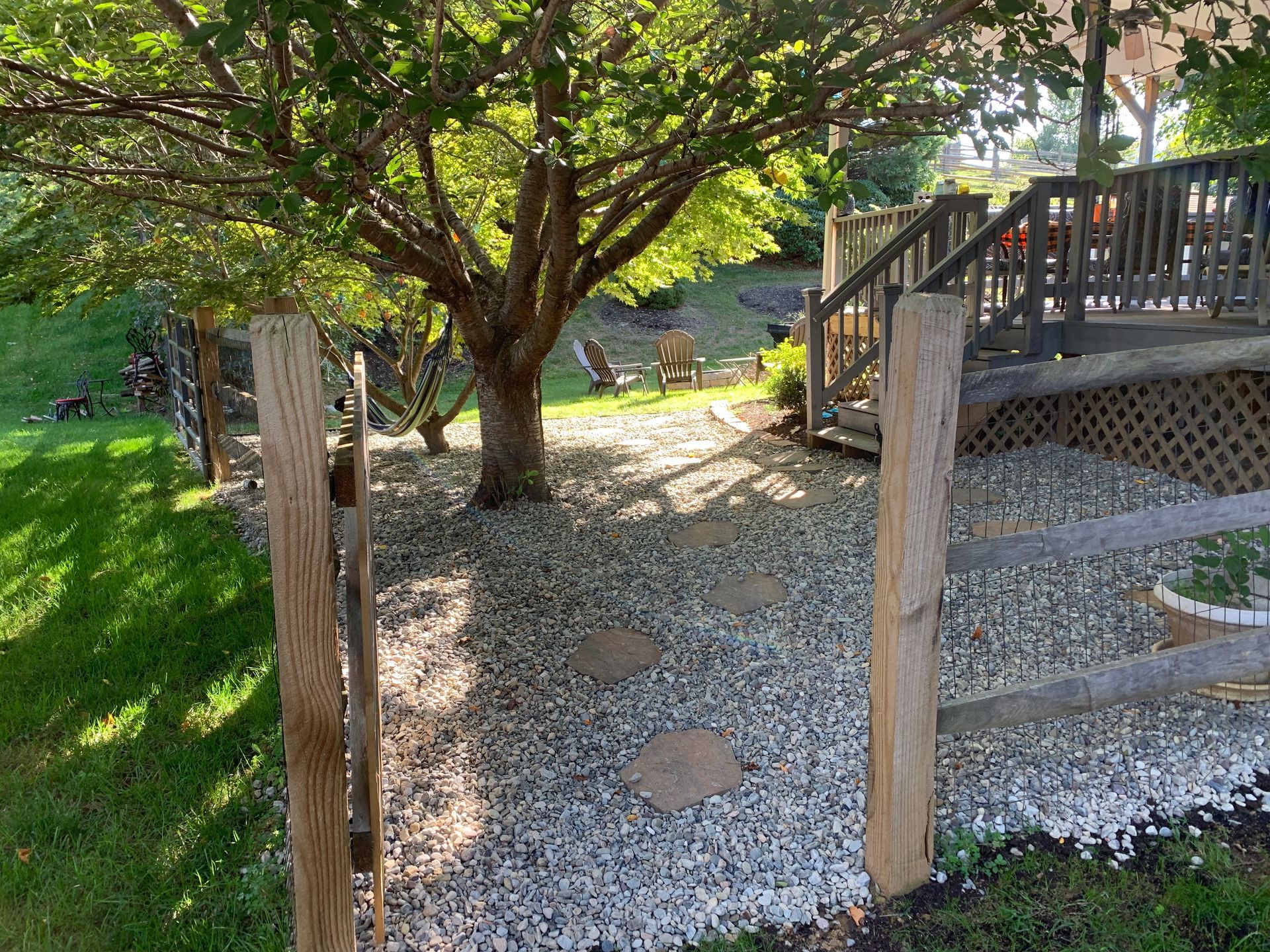 A gravel path with stone steps leads under a tree toward a wooden deck and fence in a backyard garden setting.