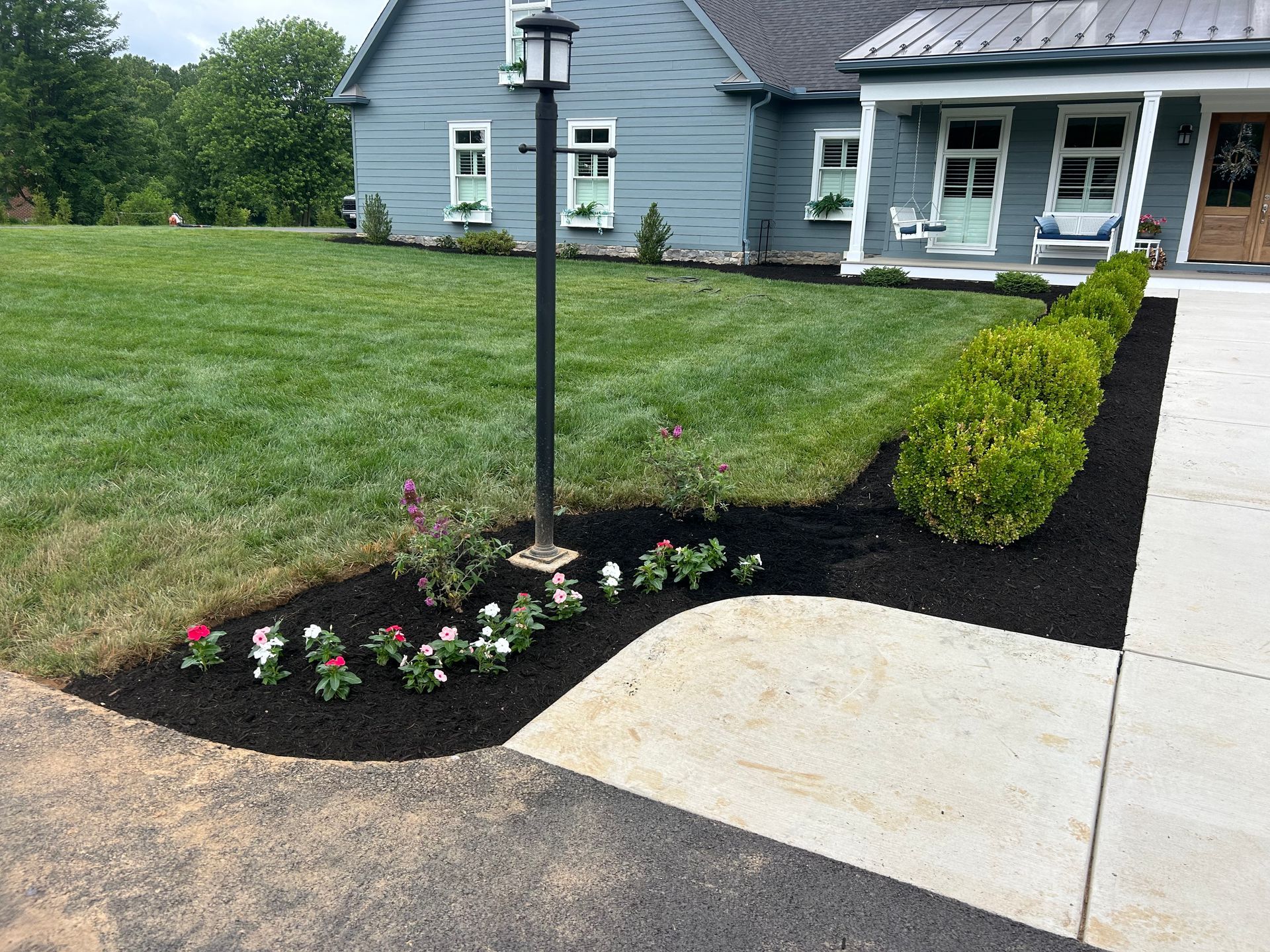 A light blue house with a porch, a green lawn, and a flower bed edged with mulch, shrubs, and flowers along a walkway.