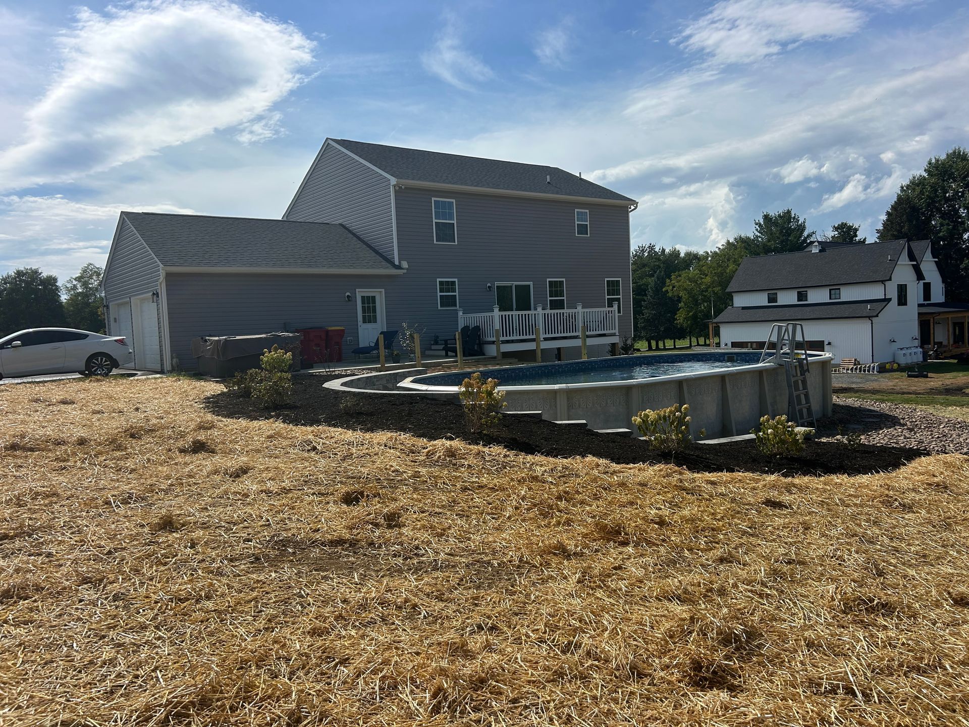 A grey two-story house with an above-ground pool and a stone retaining wall in a backyard covered in wood chips.