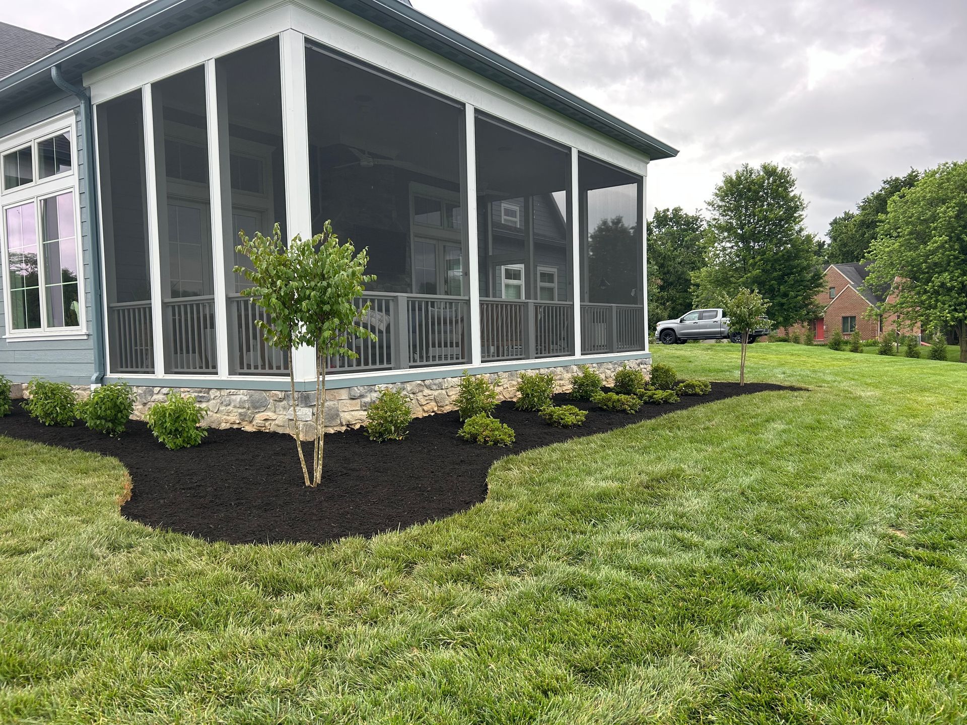 A house with a screened-in porch and stone foundation, featuring a landscaped flower bed with black mulch and small shrubs.
